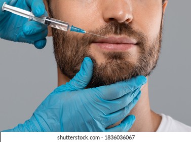 Unrecognizable Bearded Man Getting Lips Filler In Beauty Salon, Grey Studio Background, Cropped. Closeup Of Cosmetician Hands Filling Hyaluronic Acid In Man Lips. Male Cosmelogoy, Aesthetic Medicine