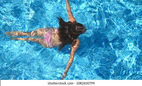 Unrecognizable beautiful girl floating across the pool of hotel. Young woman swimming in clear blue water of basin on sunny day. Concept of summer vacation or holiday. Top view Close up Slow motion.