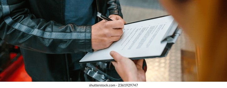 Unrecognizable biker man holding helmet signing insurance policy to receipt his repaired motorcycle on workshop while female mechanic holding the clipboard - Powered by Shutterstock