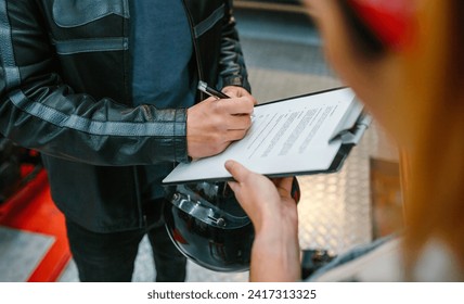 Unrecognizable biker man wearing leather jacket and holding helmet signing insurance policy to receipt his repaired motorcycle on workshop - Powered by Shutterstock