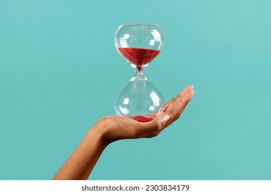 Unrecognizable black female hand holding transparent hourglass with red sand against turquoise background