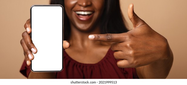 Unrecognizable black lady pointing at modern cellphone with white empty screen, mockup. Female hands holding mobile phone with blank display, copy space, panorama, isolated on beige studio background