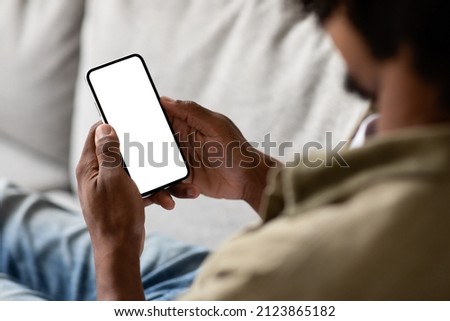 Unrecognizable Black Male Using Smartphone With Blank White Screen At Home, Over Shoulder View Of African American Guy Relaxing On Couch With Empty Mobile Phone, Mockup Image With Copy Space