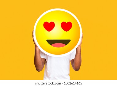 Unrecognizable Black Man Hiding Face Against In Love Emoticon Emoji, Holding Big Illustrated Smile Sticker Over Yellow Background, Copy Space