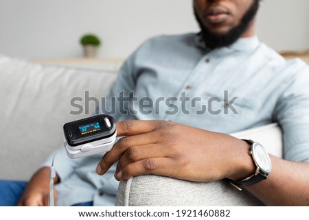 Unrecognizable Black Man With Pulse Oximeter On Hand Measuring Oxygen Saturation Level At Home. Pulseoxymeter Medical Device, Pulseoxymetry Clip Machine Monitorin Ox Rate. Cropped, Selective Focus