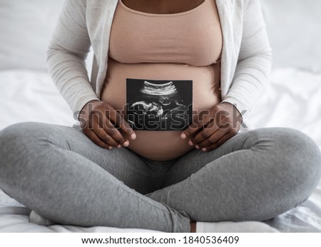 Unrecognizable black pregnant lady demonstrating her baby sonography photo while sitting on bed at home, showing firts photo of her child, enjoying happy maternity time, cropped image, closeup