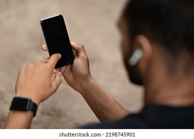 Unrecognizable Black Sportsman With Earbuds And Smartwatch Using Cell Phone With Black Blank Screen For Advertisement, Athletic Guy Having Outdoors Workout, Mockup, Closeup