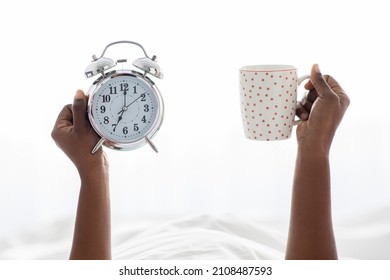 Unrecognizable Black Woman Hands Holding Cup With Strong Coffee And Alarm, Cropped Of African American Lady Waking Up In The Morning, Sitting In Bed, White Background