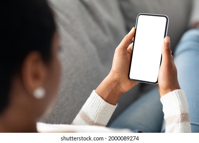 Unrecognizable Black Woman Holding Smartphone With White Screen At Home, Over Shoulder View Of African American Lady Using Blank Mobile Phone While Relaxing On Couch, Mockup Image With Copy Space