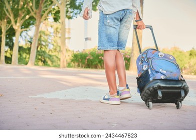 Unrecognizable boy walks through the park dragging his trolley on the way home after finishing his day of classes at school.
