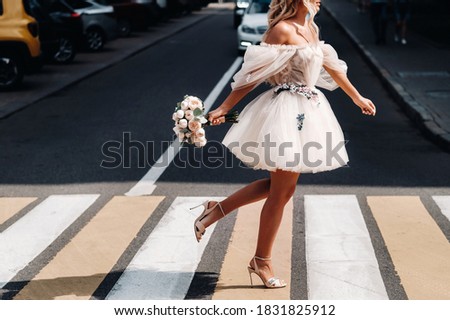 an unrecognizable bride in a short white wedding dress with a bouquet runs across the road in the city at a pedestrian crossing