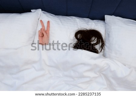 Unrecognizable brunette woman under the blanket is showing V victory hand sign. She is lying in bed on white linen. The concept of a lazy morning and unwillingness to get up. Hiding from reality.
