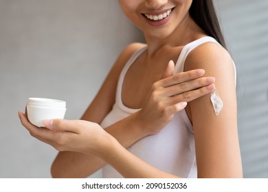 Unrecognizable brunette woman in white top applying skin butter or mosturizer on her shoulder after morning shower or bath and smiling, holding jar with cream, cropped, copy space