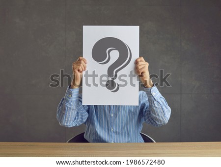 Unrecognizable business person holding paper sheet with question mark and asking questionnaire or quiz question. Faceless unknown entrepreneur sitting at desk hiding face behind question mark placard