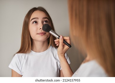 Unrecognizable caring mother applying powder with brush on face of teen girl while doing makeup against gray wall