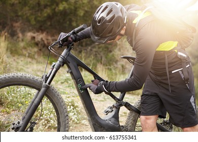 Unrecognizable Caucasian Biker In Helmet And Gloves Pressing Buttons On Frame Of His Black Electric Bike. Cropped View Of Young Rider Switching Speed Mode On Control Panel Before Cycling In Forest