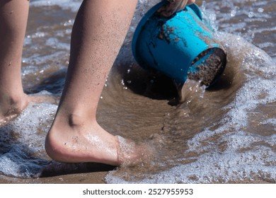 Unrecognizable Caucasian child's feet playfully interact with a blue beach bucket at the water's edge - Powered by Shutterstock