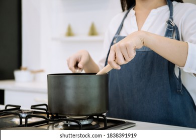 Unrecognizable Cheerful Asian woman cooking in the modern kitchen, kitchen decorated in modern and minimal style. Chief make a soup in the black pot on the gas stove. Mother prepare breakfast for kid