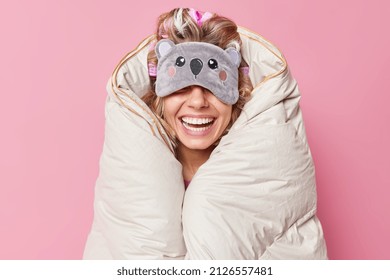 Unrecognizable cheerful woman wears sleepmask on eyes smiles broadly wrapped in duvet expresses positive emotions being in good mood isolated over pink background. People rest and bedtime concept