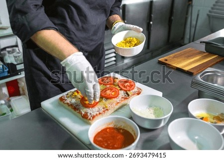 Unrecognizable chef with hygienic gloves adding corn toppings to a vegetarian focaccia in restaurant. Preparation of fresh and organic pizza on stainless steel counter on professional kitchen.