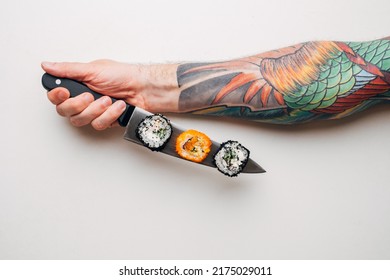 An Unrecognizable Chef Man With A Tattoo On His Arm Holds Sushi Rolls On A Knife On A White Background.