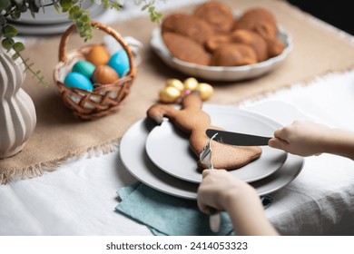 Unrecognizable child cutting gingerbread cookie shaped as Easter bunny with fork and table knife at festive decorated table. Holiday traditions, learning table manners and dining etiquette