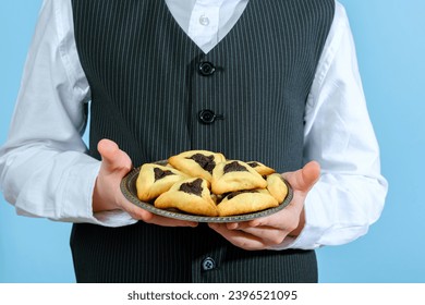 An unrecognizable child is holding a plate of festive food Hamantaschen cookies with poppy seed filling on the blue background. Purim celebration concept. Jewish carnival holiday. - Shutterstock ID 2396521095