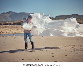 Unrecognizable child standing alone on sandy beach in sunny windy day and looking away while wrapped in white sheet