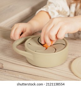 Unrecognizable child taking piece of carrot out from pastel gray silicone snack cup near lid at wooden table. Baby accessories, tableware, first feeding concept. Top view, instagram use, square frame.