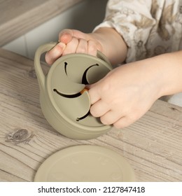 Unrecognizable child taking piece of snack from pastel gray silicone snack cup near lid at wooden table. Baby accessories, tableware, first feeding concept. Top view, instagram use, square frame.