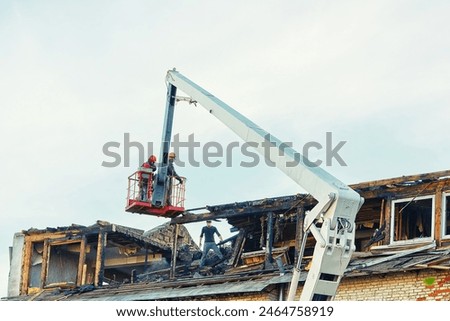 Unrecognizable  construction team operating crane to dismantle charred roof in the aftermath of fire. Post-fire dismantling of a burnt roof with a crane cradle and team of workers