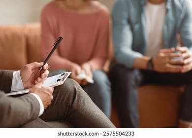 Unrecognizable couple having therapy session at psychotherapists office, selective focus on man professional psychologist holding pen and clipboard, writing in notepad, talking and consulting clients