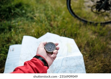 Unrecognizable cyclist using compass over map. A hand is holding the compass, the floor is grass and we can see part of bike wheel. Orientation in adventure concept.