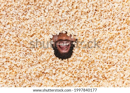 Unrecognizable dark skinned bearded man hidden in heap of salty popcorn. Male mouth with broad smile surrounded by air popped corn. Funny guy buried in delicious snack. Food and people concept