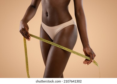 Unrecognizable Dark Skinned Woman African American Model In Nude Panties Holding Measuring Tape Over Her Sexy Smooth Legs, Posing On Beige Studio Background. Thigh Slimming Concept