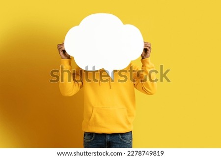 Unrecognizable dark-skinned man in casual outfit hiding his face head behind empty white communication bubble with free space for text or image, yellow studio background
