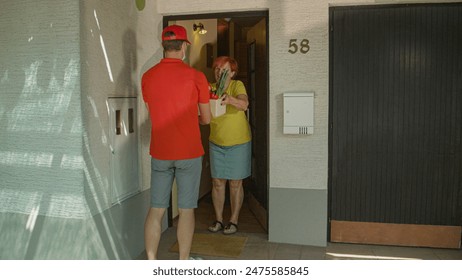 Unrecognizable delivery guy wearing surgical mask hands elderly woman her box full of organic groceries. Young male courier delivers a parcel of fresh produce to senior lady during covid19 lockdown. - Powered by Shutterstock