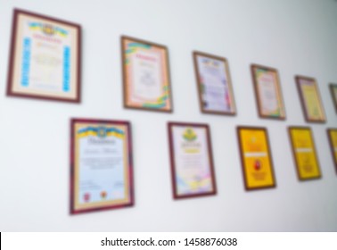 Unrecognizable Diplomas, Certificates And Awards In Wooden Frames On The White Office Wall. Abstract Blurry Image.