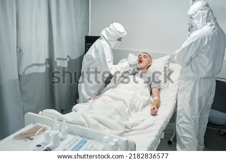 Unrecognizable doctors wearing white protective suits with masks testing mature man for infectious or bacterial disease