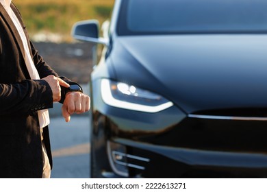 Unrecognizable elegant man using smartwatch app for remote controlling of premium class electric car, driver distantly approaching car and opening door, smart car.