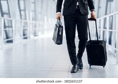 Unrecognizable entrepreneur in black suit walking by airport terminal, carrying luggage and bag, copy space. Rich businessman with case and suitcase going to business trip, cropped - Powered by Shutterstock