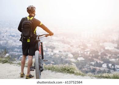 Unrecognizable European male cyclist relaxing on top of mountain, keeping hands on handlebar of his electric pedal-assist bicycle, enjoying amazing urban landscape below. Travel and active lifestyle