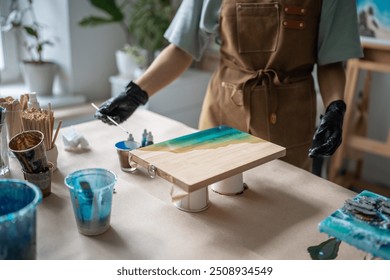 Unrecognizable female artist applies paint on epoxy with long stick, designing pattern for future artwork on wooden board in workshop. Creative occupation, art idea, small business, do it yourself - Powered by Shutterstock