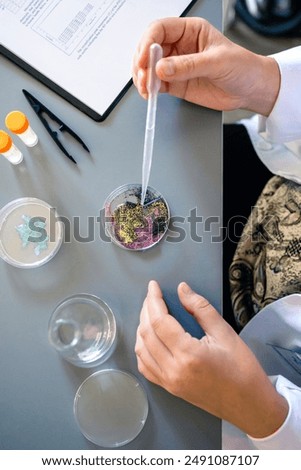 Unrecognizable female chemist hands adding drop of liquid from from pipette over petri dish with mix of colorful glitters in environment research lab. Study of microplastics composition concept.
