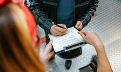 Unrecognizable Female Mechanic With Red Hair Bandana Holding A Clipboard While Biker Man Wearing Leather Jacket Signing Insurance Policy To Receipt His Repaired Motorcycle