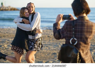 Unrecognizable female photographing cheerful multiracial female friends embracing each other while standing on sandy beach near rippling sea on sunny day