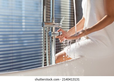 Unrecognizable Female Playing With Water Flowing From Tap While Sitting On Bathtub Edge In Luxury Bathroom, Woman Wrapped In Towel Ready To Take Bath At Home, Adjusting Temperature With Hand, Cropped