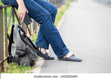 Unrecognizable Female Sitting On Steel Fence Of Pedestrian Pathway With Mobile Phone In Hand, Urban Backpack Standing On Earth 