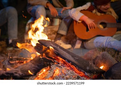Unrecognizable friends having fun playing music and enjoying bonfire camping in nature at night. Group of people chilling at fire in the evening, camping in the forest near lake. High quality photo