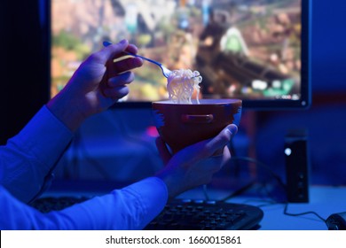 Unrecognizable Gamer Guy Eating Noodles Sitting In Front Of Computer And Playing Shooter Game Indoor At Night. Closeup, Selective Focus